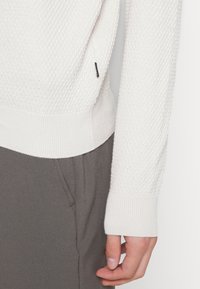 Lys beige strikkegenser med et strukturert mønster, ribbede mansjetter og nederkant, kombinert med grå bukser. Subtil merkevare på siden.