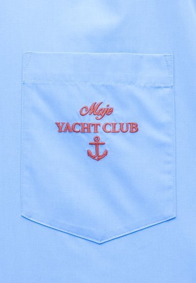 Φωτεινή γαλάζια υφασμάτινη τσέπη με κόκκινη κεντημένη επιγραφή "Maje Yacht Club" και σύμβολο άγκυρας κάτω από το κείμενο.