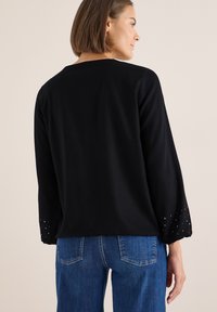 Schwarzes Oberteil mit langen Ärmeln, das mit Nietenakzenten an den Manschetten versehen ist. Glattes Gewebe mit klassischem Schnitt, kombiniert mit blauen Jeans.