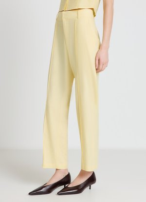 Donna che indossa pantaloni gialli a vita alta con gamba larga abbinati a scarpe nere con tacco basso e punta stretta, in piedi davanti a uno sfondo semplice.