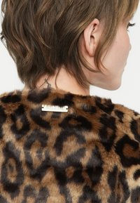 Cabello corto castaño y una chaqueta de piel sintética de leopardo con una etiqueta metálica visible en la parte posterior del cuello, usada por una persona de perfil.
