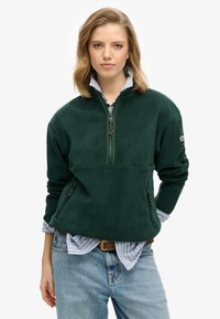 Groene fleece pullover met een ritskraag, zijzakken en een zachte textuur, over een blauw-wit gestreept shirt.