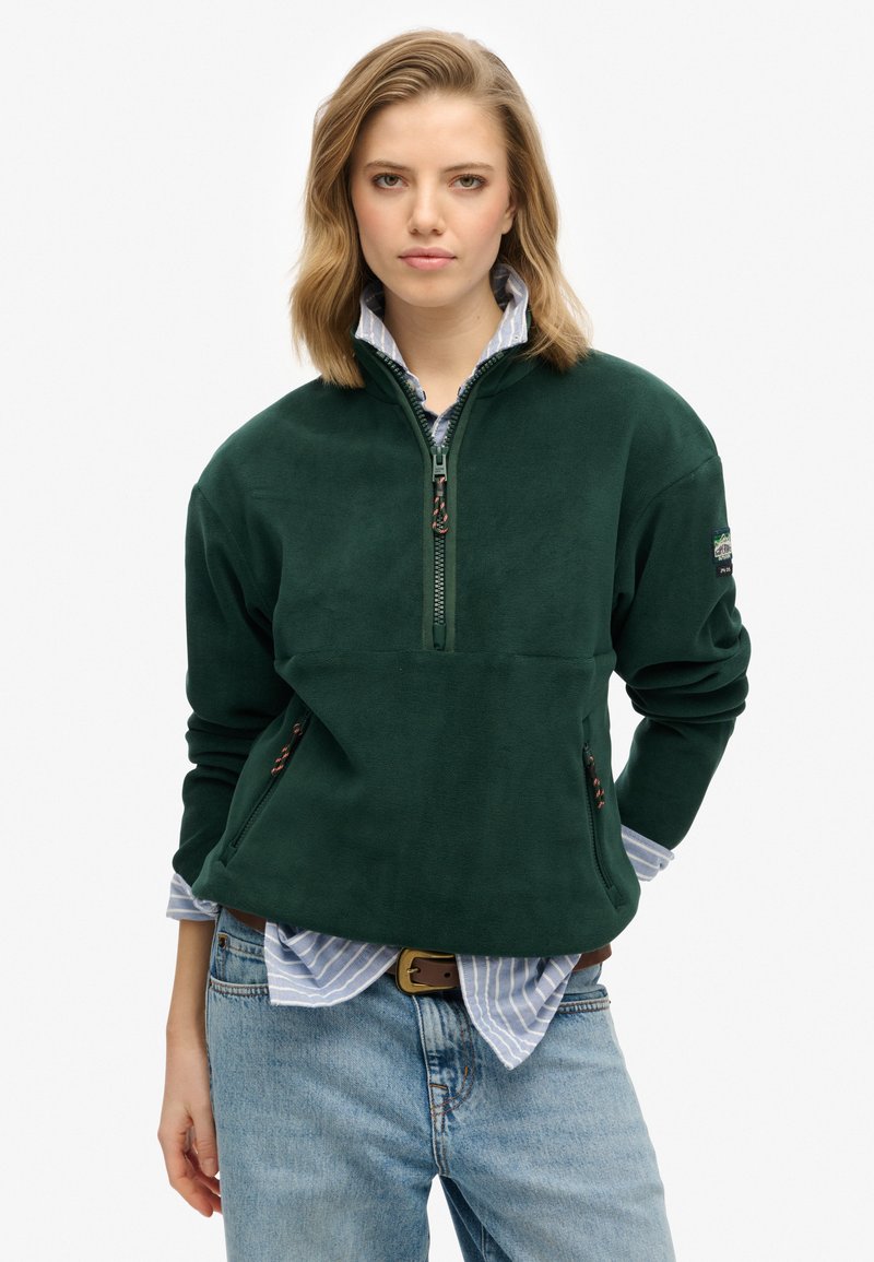 Groene fleece pullover met een ritskraag, zijzakken en een zachte textuur, over een blauw-wit gestreept shirt.