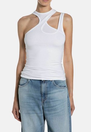 Donna che indossa un top bianco asimmetrico senza maniche con ritagli abbinato a jeans a vita alta azzurri su uno sfondo semplice.