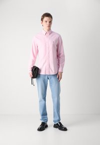 Gaiši rozā krekls ar pogām, gaiši zili džinsi, melnas kurpes, tur rokā maza melna crossbody soma. Minimālisma dizains, ikdienas piegriezums.