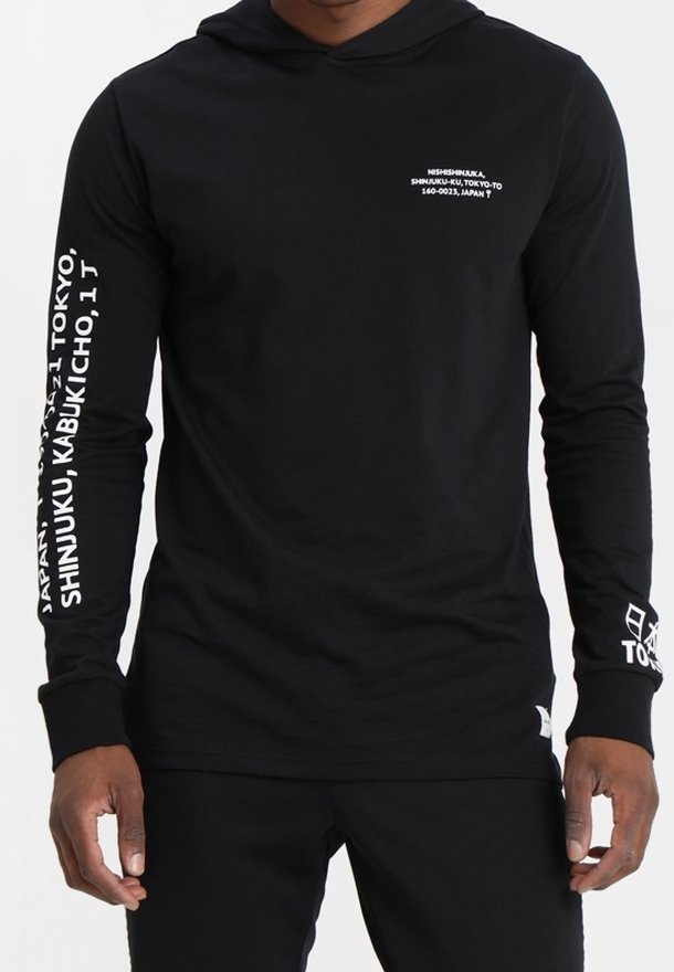 Hoodie noir à manches longues avec texte en japonais et en anglais blanc sur la poitrine et les manches, porté par une personne debout.