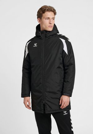 Homme portant une veste de sport noire à capuche avec des accents blancs aux épaules et un pantalon de sport noir sur un fond clair uni.