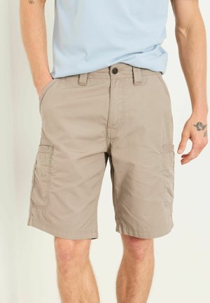 Homme portant un short cargo marron clair et une chemise bleu clair, mains dans les poches, montrant les détails des poches avant et latérales.
