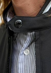 Zwarte jas met een hoge kraag, voorzien van een glanzende zwarte knoop en een gestreept wit en blauw shirt eronder. Stijlvolle stoftextuur.