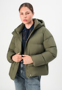 Jonge vrouw in een olijfgroene pufferjack met een capuchon, een marineblauw kanten top en vervaagde high-waisted jeans tegen een witte achtergrond.