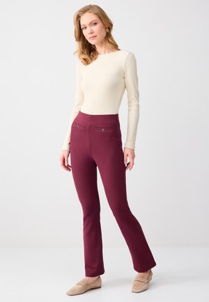 Vrouw staand, gekleed in een crèmekleurige top met lange mouwen, wijnrood hoge taille broek met zakken met knopen, en beige loafers tegen een effen achtergrond.