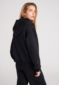 Sudadera con capucha negra de corte relajado, con puños y dobladillo acanalados. Presenta una textura suave y un diseño minimalista.