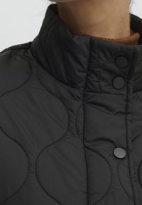 Chaqueta acolchada negra con cuello alto, con botones redondos negros y un patrón texturizado de líneas curvas. Material suave y ligero.