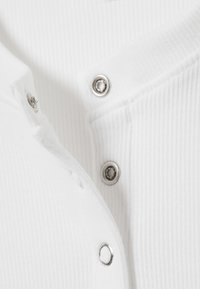 Tissu blanc côtelé avec une patte de boutonnage dotée de pressions argentées. La texture est lisse, avec une légère élasticité et un design épuré et minimaliste.