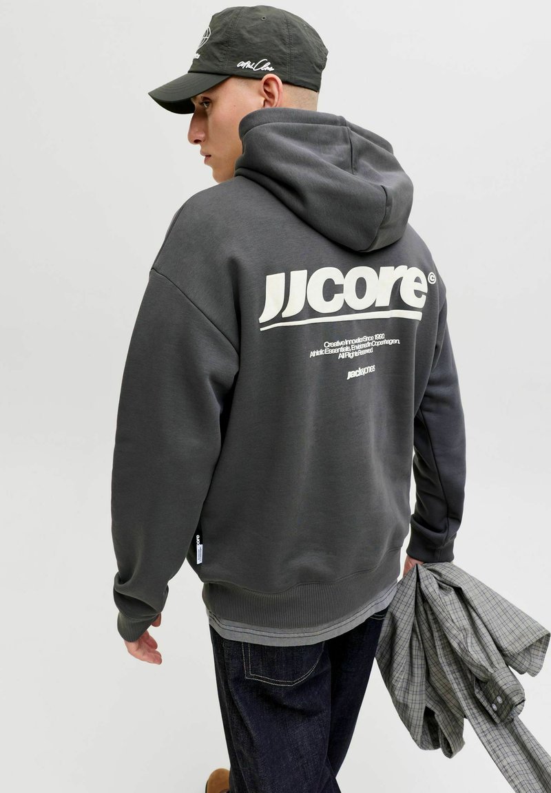 Ung mand iført en mørkegrå JJ Core hættetrøje og sort kasket, der holder en grå ternet skjorte i venstre hånd og vender let væk til siden.