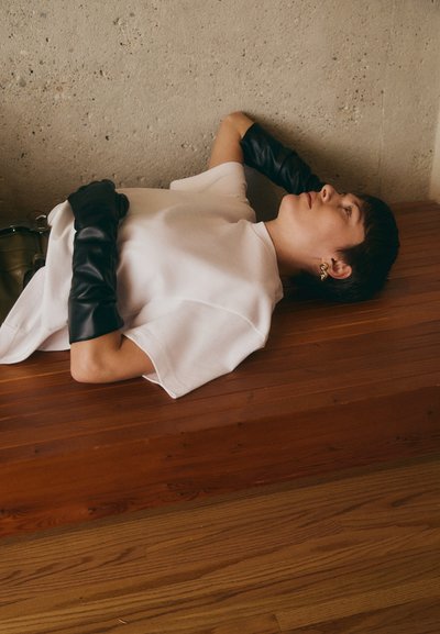 Λευκό oversized t-shirt σε συνδυασμό με μακριά μαύρα δερμάτινα γάντια, τοποθετημένο σε μια ξύλινη επιφάνεια μπροστά σε έναν υφασμάτινο τοίχο από σκυρόδεμα.