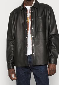 Giacca nera in pelle con bottoni a pressione, taschino sul petto con la scritta "YPS" in rilievo e una texture morbida e liscia. Indossata sopra una camicia a fantasia.