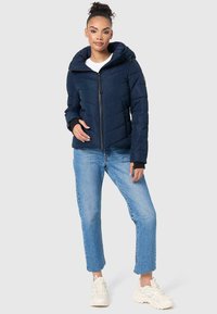 Veste matelassée en duvet marine avec capuche, accents noirs, fermeture éclair frontale et poignets élastiques. Associée à un jean bleu clair et des baskets blanches.