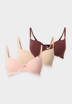 Femme debout pieds nus sur fond blanc, portant un soutien-gorge marron foncé assorti et une culotte bordée de dentelle.