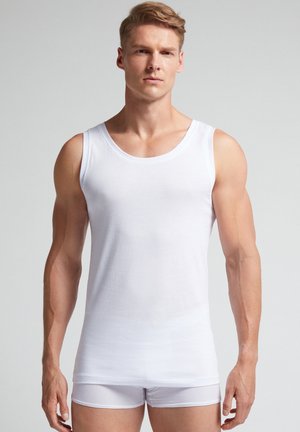 Weißes Baumwoll-Top mit Rundhalsausschnitt und weiten Armöffnungen, kombiniert mit weißen Boxershorts, Modell steht vor einem hellgrauen Hintergrund.