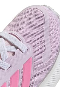 Zapatilla deportiva de color púrpura claro con tejido de malla, acentos en rosa y cordones blancos; presenta una suela texturizada y elementos de diseño distintivos.