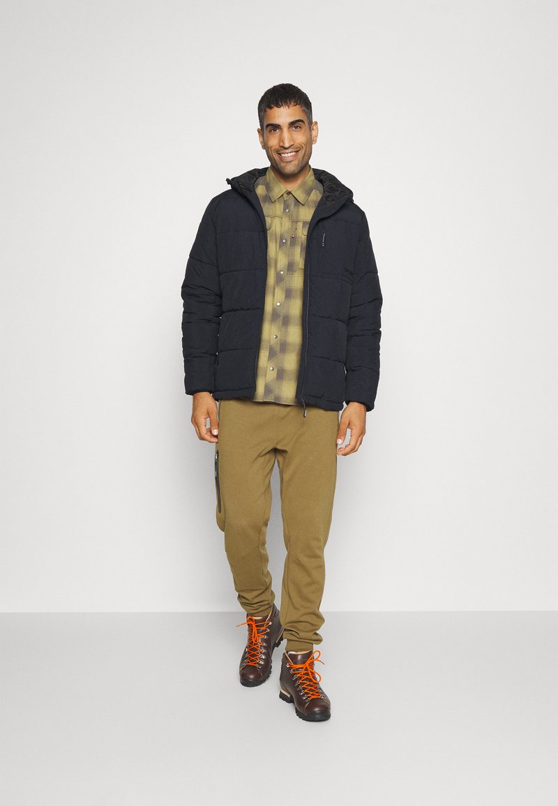 Giacca a vento nera con cappuccio, camicia a quadri oliva, joggers khaki e stivali marroni con lacci arancioni. Presenta cerniere sui pantaloni.