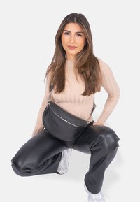 Schwarze Leder-Umhängetasche mit geschwungener Form, silbernen Reißverschlussdetails und verstellbarem Gurt, getragen über einem gerippten beigen Oberteil und schwarzen Hosen.