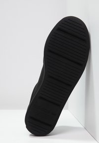 Černá podrážka tenisky Adidas s horizontálními drážkami a texturovaným gripem, umístěná vertikálně proti bílým rohovým stěnám.