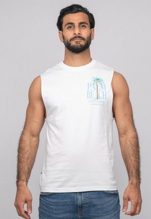 Homme barbu portant un débardeur blanc avec un motif de palmier et le texte "Palm Beach", assorti à un jean bleu, debout devant un fond gris clair.