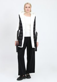 Gilet puffa nero con rivestimento in finta pelliccia bianca, caratterizzato da un design lungo, bottoni a pressione, tasche laterali e dettagli neri. Indossato sopra un maglione bianco e pantaloni neri.