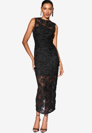 Abito nero senza maniche con design in pizzo floreale, silhouette aderente, parte inferiore trasparente e orlo al ginocchio. Indossato con tacchi neri a strisce.