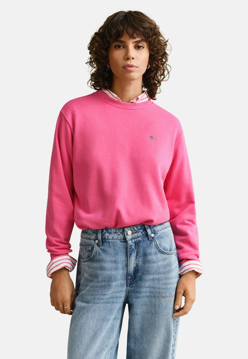 Jeune personne aux cheveux bouclés portant un sweat-shirt rose vif par-dessus une chemise à rayures et un jean taille haute bleu clair, debout sur un fond blanc.