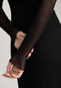 Schwarzes Kleid mit langen, transparenten, eng anliegenden Ärmeln, die die Haut darunter offenbaren. Die Textur ist glatt, mit einem subtilen gerippten Muster an den Ärmelbündchen.