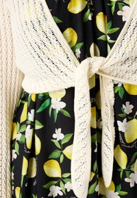 Gilet crème en tricot noué à la taille sur une robe noire avec un motif de citrons et de fleurs, mettant en valeur des citrons jaunes vifs et des feuilles vertes.