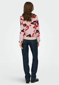 Pull en tricot présentant un motif floral abstrait coloré sur fond rose clair, avec des poignets côtelés et un ourlet crème. Associé à un jean noir.