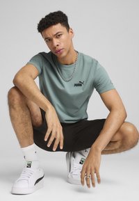 Světle modré tričko s černým logem Puma, zkombinované s černými šortkami a bílými teniskami. Model klečí, má krátké hnědé vlasy a řetízkový náhrdelník.