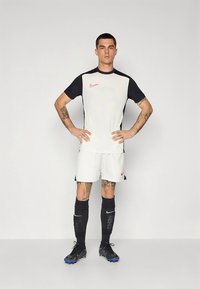 Balta krekla ar melnām īsām piedurknēm, uz kuras atrodas sarkans Nike logotips, kopā ar baltām šortām, melnām zeķēm un melnu apavu ar ziliem akcentiem.