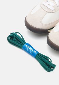 Adidas gröna skosnören på en grå och krämfärgad sneaker, med texturerade mocka- och nylondetaljer, samt vita öljetter och snören.