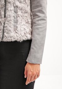 Main d'une femme reposant sur le côté, portant une veste en fausse fourrure grise aux manches grises lisses et une jupe noire.