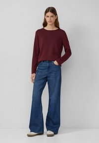 Bordeauxfarbenes langärmliges Oberteil, kombiniert mit weit geschnittenen blauen Jeans. Das Oberteil hat einen runden Halsausschnitt und eine glatte Textur, während die Jeans eine hohe Taille haben.