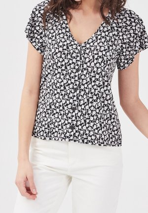 Femme portant un chemisier noir à boutons avec un imprimé floral blanc et des manches courtes, associé à un pantalon blanc sur un fond uni.