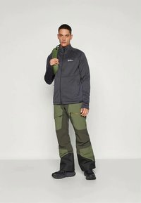 Moška temno siva jakna z zadrgo in teksturirano površino, parjena z zelenimi in temno sivimi hlačami v barvnih blokih. Črne čevlje dopolnjujejo videz.