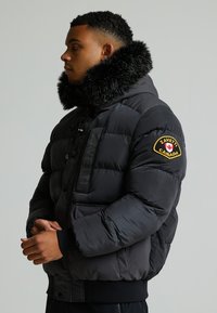 Čierna puffer bunda s golierom z umelej kožušiny, zapínaním na zips a gombíky, nášivkou loga na rukáve a rebrovanými manžetami. Textúrovaný, teplý a izolovaný dizajn.