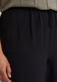 Černé texturované kalhoty širokých nohavic s elastickým pasem a šňůrkou na stažení. Detailní pohled na pas a ruku odpočívající na látce.