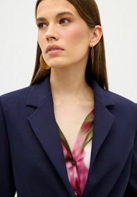 Námorně modrý blejzr s klopami, nošený přes hedvábnou květinovou blůzu se vzory v růžové a zelené barvě. Viditelné zlaté kruhové náušnice.