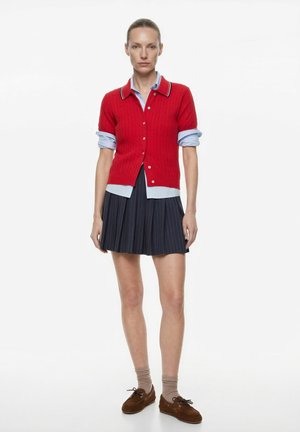 Kvinde står med armene afslappet, iført en rød kortærmet cardigan over en blå skjorte, navy plisseret nederdel, beige sokker og brune sko.