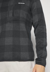 Vue mi-corps d'une personne portant un pull Columbia à carreaux gris foncé et un pantalon de couleur claire, avec le logo Columbia visible sur la poitrine.