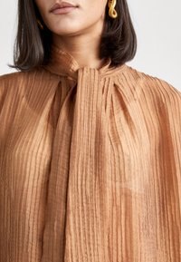 Donna che indossa una blusa plissettata di un marrone chiaro, con un colletto a fiocco annodato e grandi orecchini ad anello dorati, mostrata dalla gola al petto.