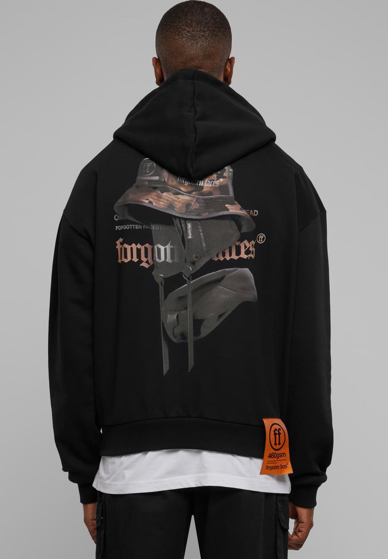 Fekete kapucnis pulóver, hátulján terepmintás sapkát ábrázoló grafikus nyomat, látható szöveggel és narancssárga címkével az alján. Puha anyag, laza szabás.