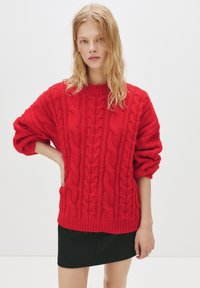 Pulover roșu tricotat, cu un model de cablu gros, guler rotund și mâneci supradimensionate, asortat cu o fustă neagră ajustată.
